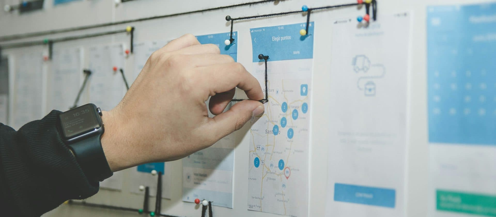 A hand pinning string between printed screens on a wall, mapping how one step of a process connects to the next.